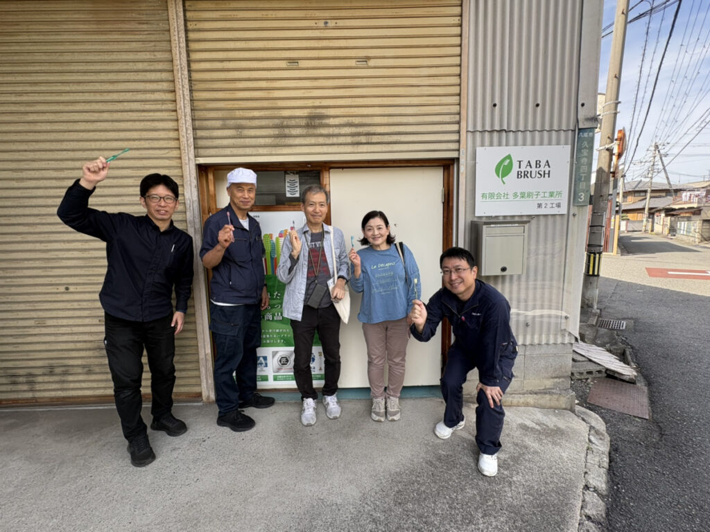 第2工場のシャッター前で、5人が色とりどりの歯ブラシを手に並び、笑顔で写る集合写真