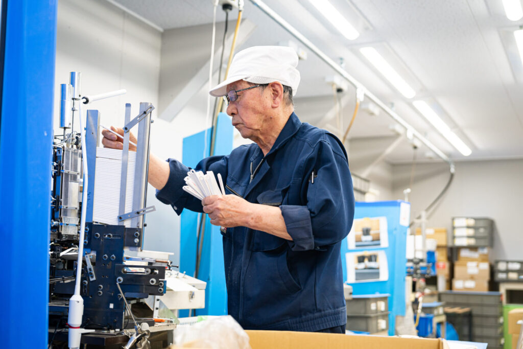 多葉刷子工業所の職人が、金属レールの精密機械の前で歯ブラシの柄を扇状に持ち検品。職人の手仕事と目利きが光る製造工程
